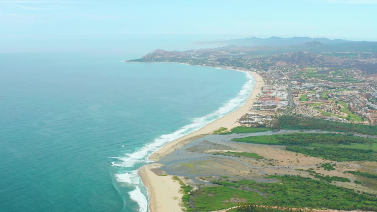 A high aerial view over La Playita Los Cabos shows turquoise ocean, a curved sandy beach, coastal buildings, green vegetation, and distant hills with no visible people.
