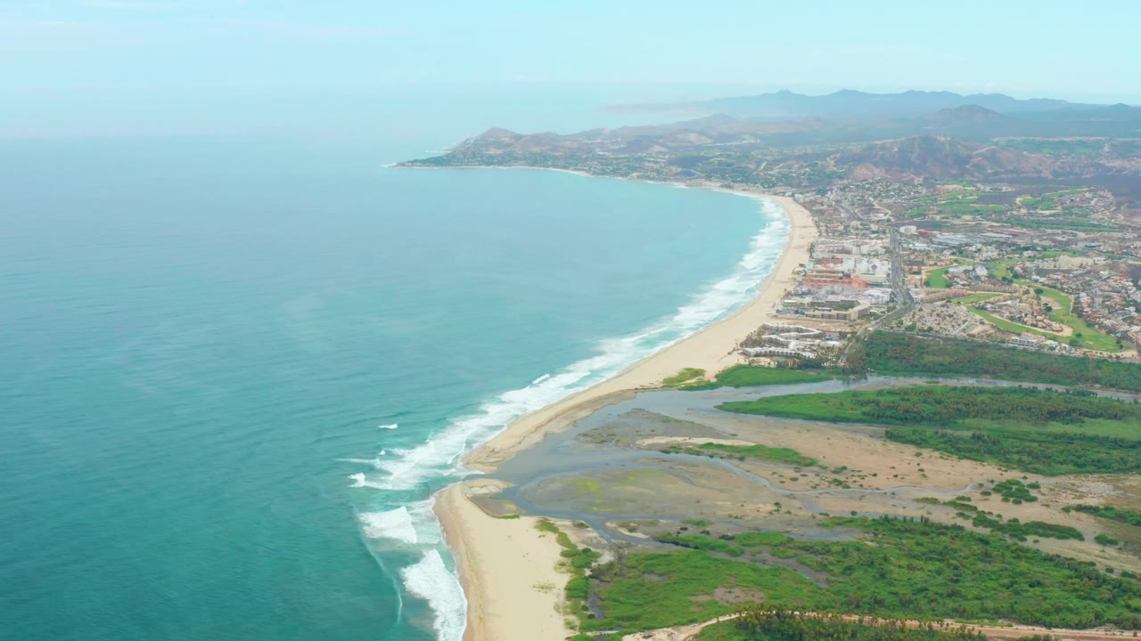 Aerial beach and coastal settlement with surf, lagoon flats, and hills beyond.