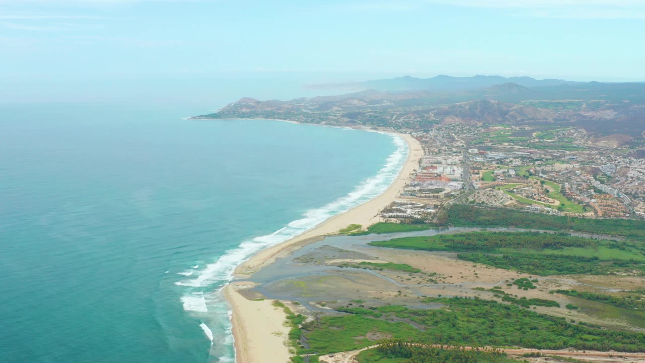 Opening aerial frame over La Playita Los Cabos, Mexico, with turquoise ocean at left, a long sandy beach, green shoreline, scattered buildings, distant mountains, clear sky, and no visible people.