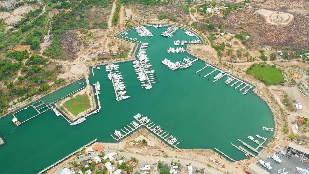 One second into the clip, the La Playita Los Cabos harbor remains centered with clustered white boats along slips, open green water to the right, dry hillside terrain above, and no visible people.