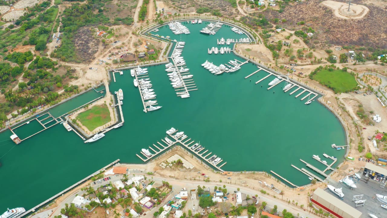 Aerial frame of La Playita Los Cabos marina with the central dock lines and moored boats prominent, dry hills and shoreline buildings around the basin, and no visible people.