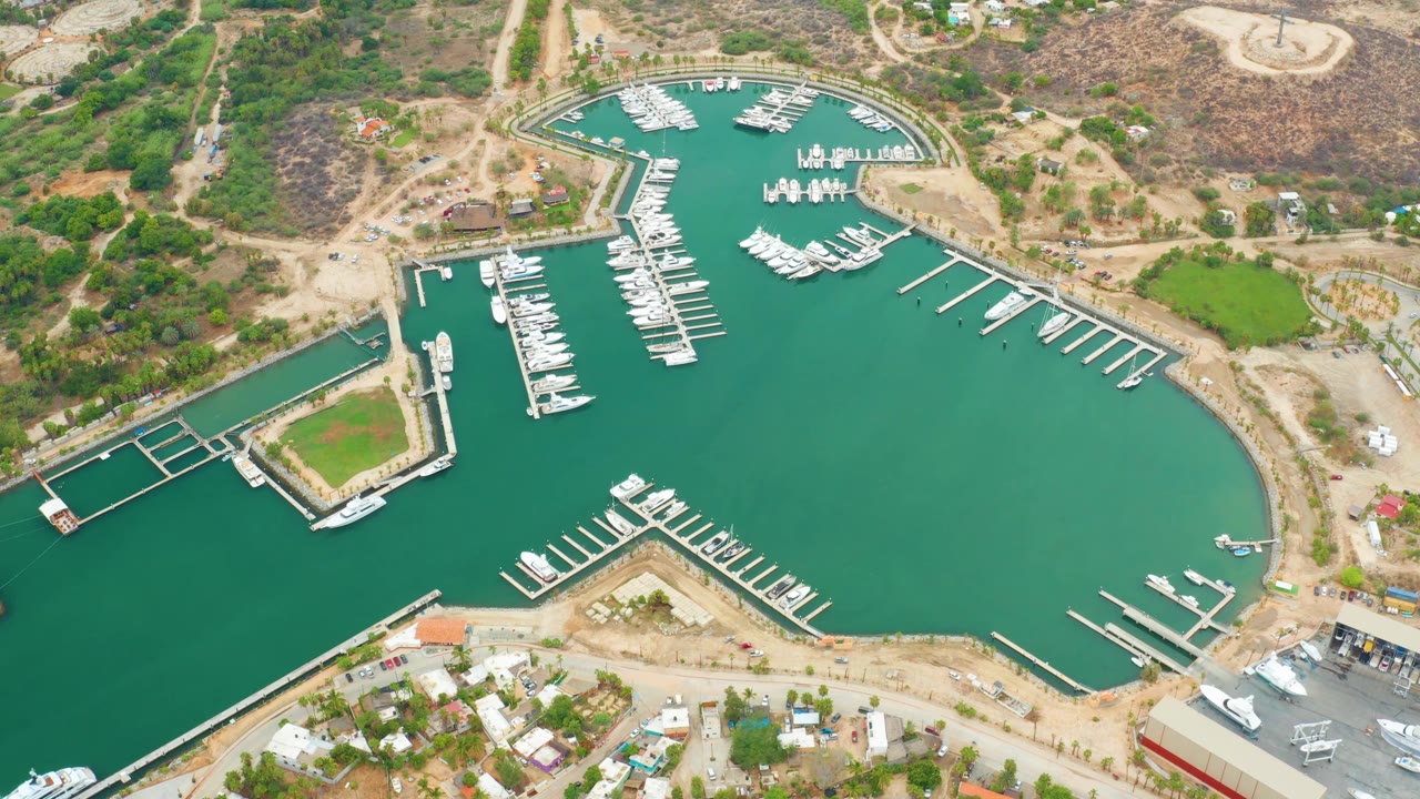 High aerial frame over La Playita Los Cabos with green-blue marina water filling the right side, clustered white boats near the top docks, hillside tracks, buildings, and no visible people.