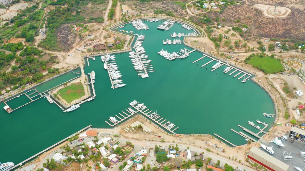 Aerial view of La Playita Los Cabos marina with white slips crossing green water, clustered boats near the upper basin, hillside roads, small buildings, and no visible people.