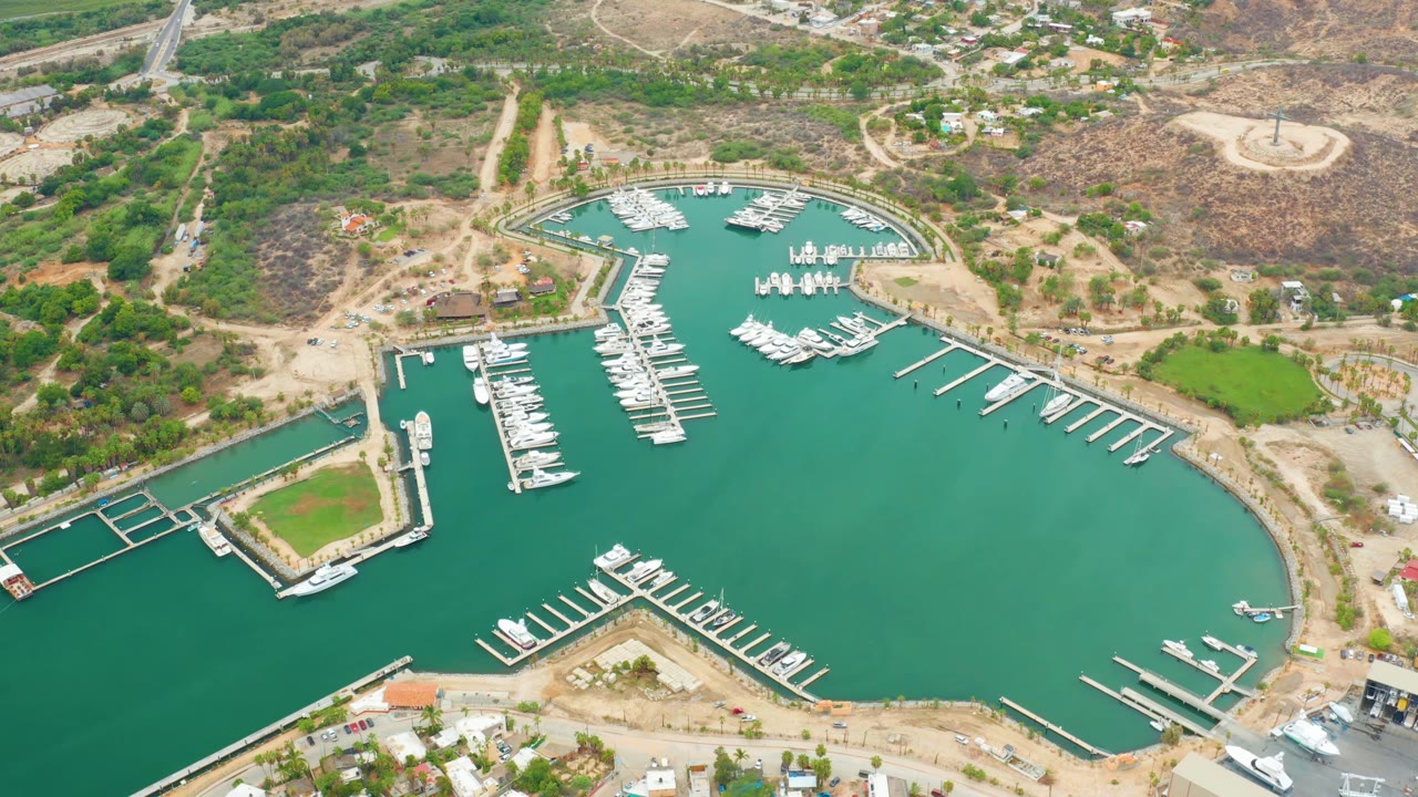 High aerial frame of La Playita Los Cabos marina, showing docked boats in green water, a rectangular waterfront parcel, piers, dry hillside roads, and no visible people.