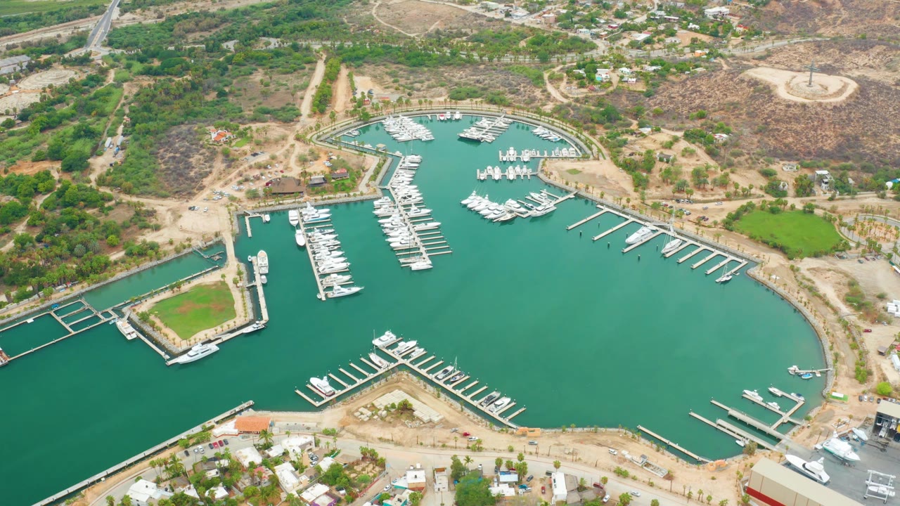 Aerial frame over La Playita Los Cabos showing the marina basin centered in green water, rows of white docks, moored boats, sandy banks, hillside roads, and no visible people.