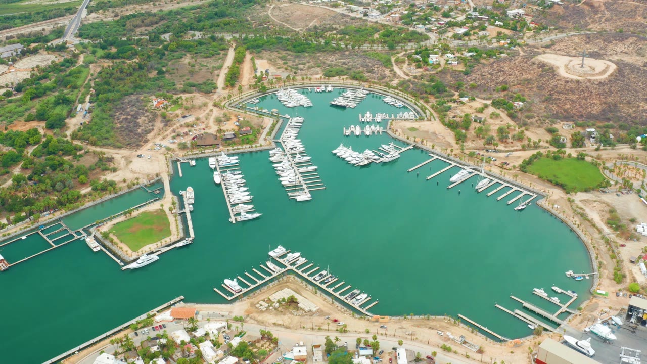 Mid-clip aerial frame over La Playita Los Cabos showing long white docks, clustered boats, green marina water, nearby low buildings, sandy tracks, dry hills, and no visible people.
