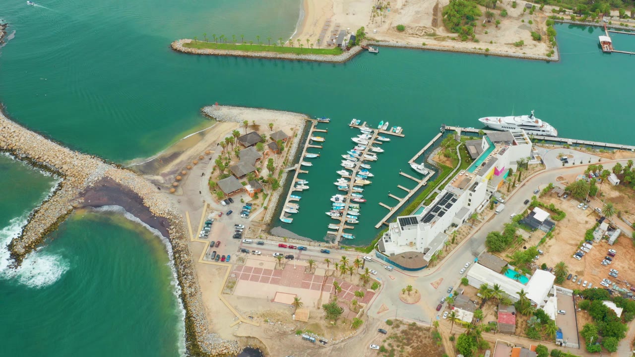 Aerial frame over La Playita Los Cabos showing green water around a rocky breakwater, marina docks, a large yacht near the quay, sandy lots, parking areas, and no visible people.