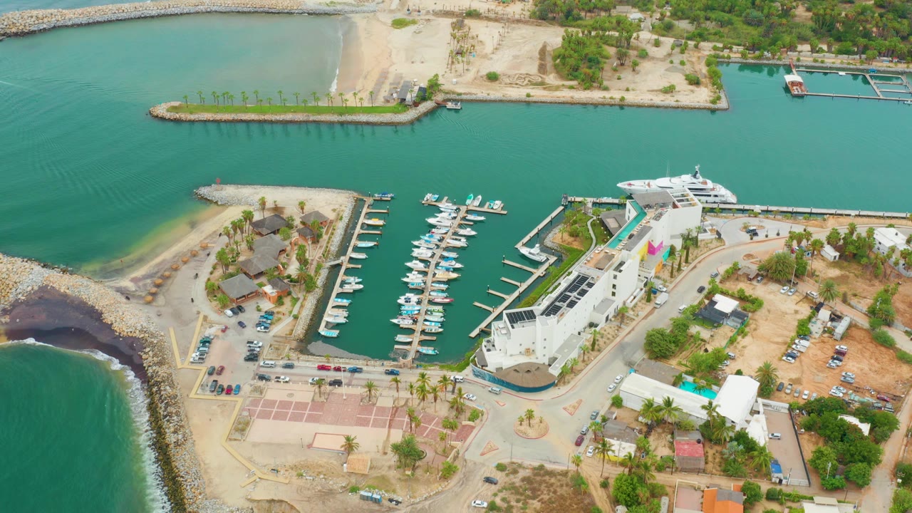 Aerial frame over La Playita Los Cabos showing turquoise water, a rocky breakwater, docked boats, a white waterfront building, sandy roads, palms, parked cars, and no visible people.