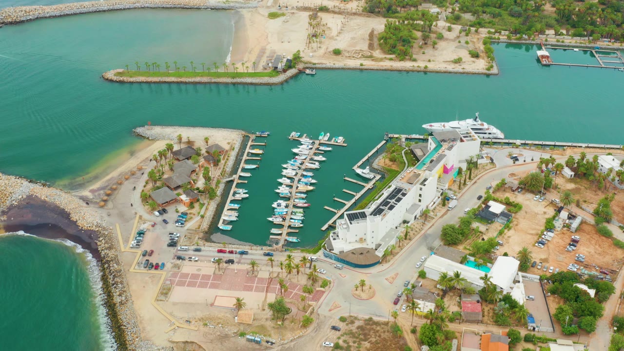 Aerial view of La Playita Los Cabos marina one second in, with green-blue water surrounding the breakwater, boats lined along docks, waterfront buildings, palms, roads, and no visible people.