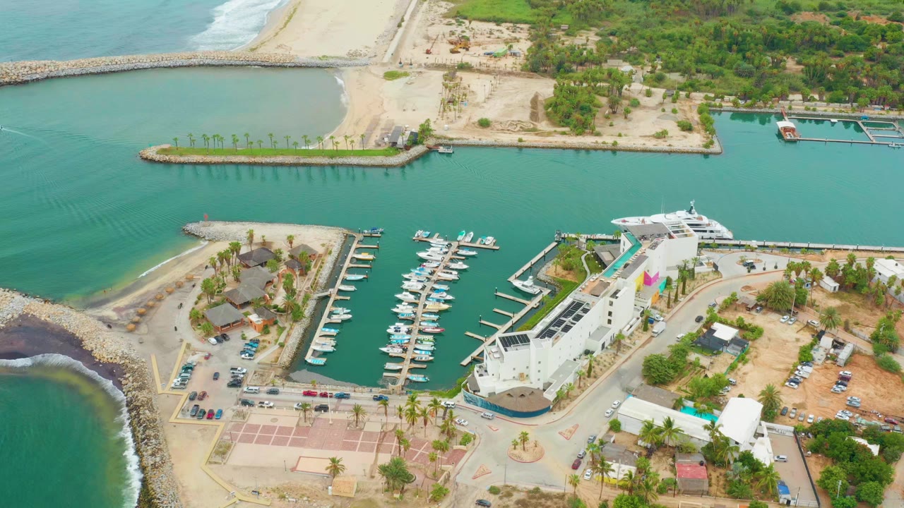 Aerial view of the La Playita Los Cabos marina with the dock rows centered, white boats moored in green-blue water, beach and brush beyond, and no visible people.