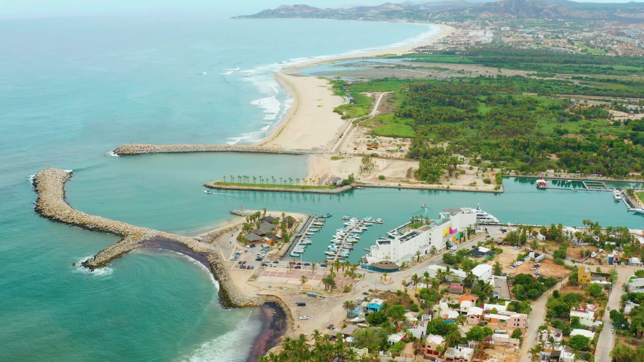 Opening aerial frame over La Playita Los Cabos showing a curved stone breakwater, marina slips, sandy beach, green shoreline, distant hills, and no visible people.