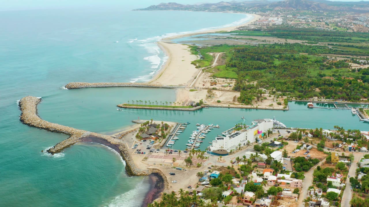 Aerial view of La Playita Los Cabos with the harbor channel centered, boats docked below, the pale beach extending into the distance, and no visible people.
