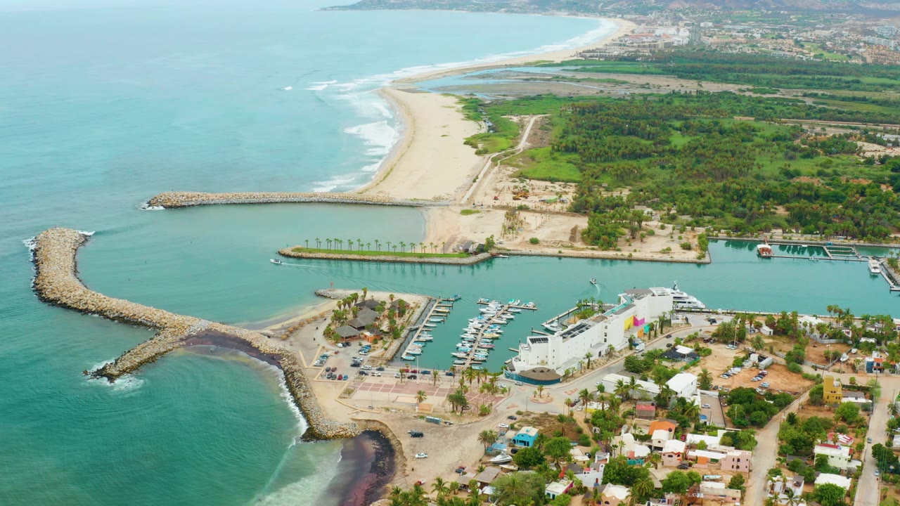 Drone view of waves near a breakwater with marina, beach, and coastal greenery behind.