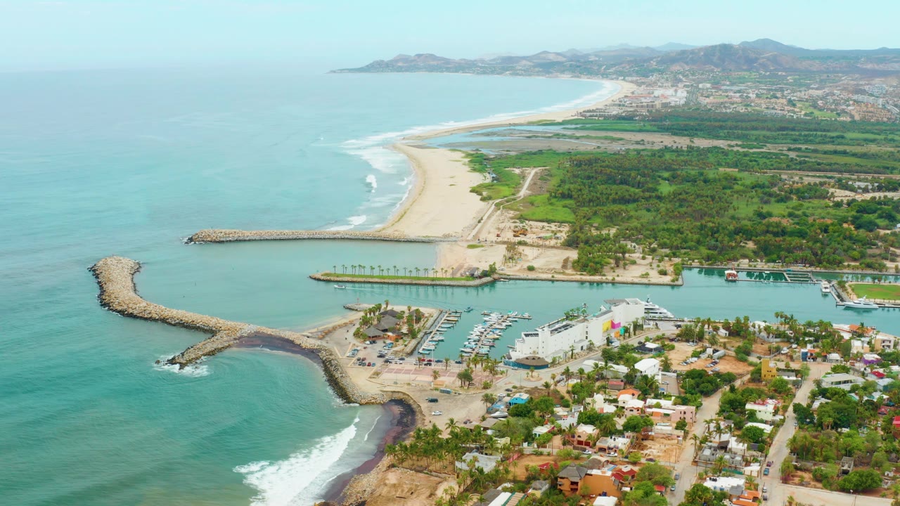 Aerial view over La Playita Los Cabos showing turquoise ocean, rocky breakwaters, a marina with boats, sandy beach, green land, distant hills, and no visible people.