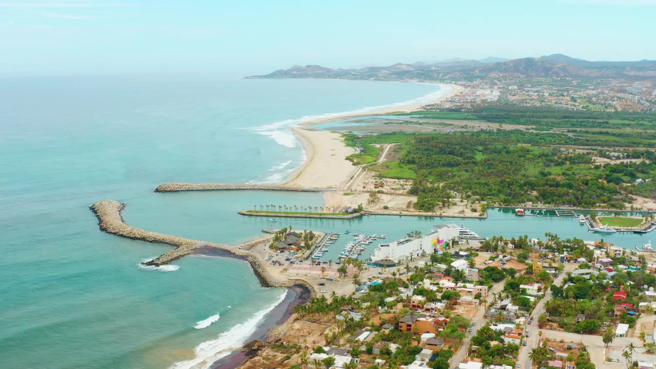 A bright aerial frame shows La Playita Los Cabos from above, with open water, a protected harbor, visible docks and boats, shoreline homes or buildings, green lots, distant hills, and no visible people.