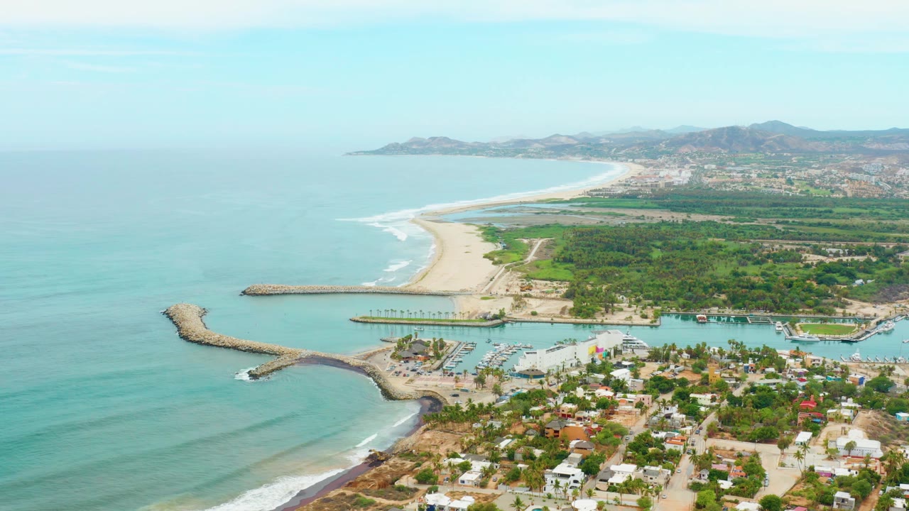 Wide aerial frame of La Playita Los Cabos with the harbor entrance below center, pale beach and surf line extending rightward, low houses in the foreground, and no visible people.