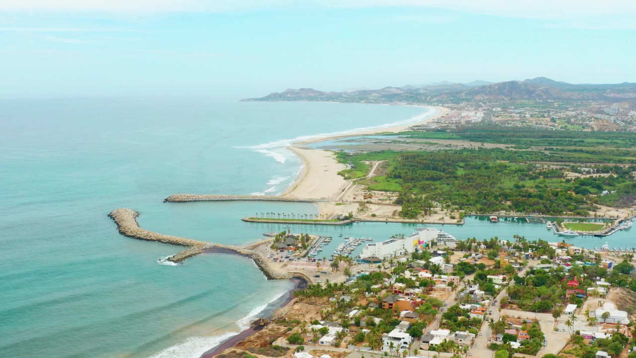 Aerial coastline with ocean, curved breakwater, small harbor, houses, and green shore.