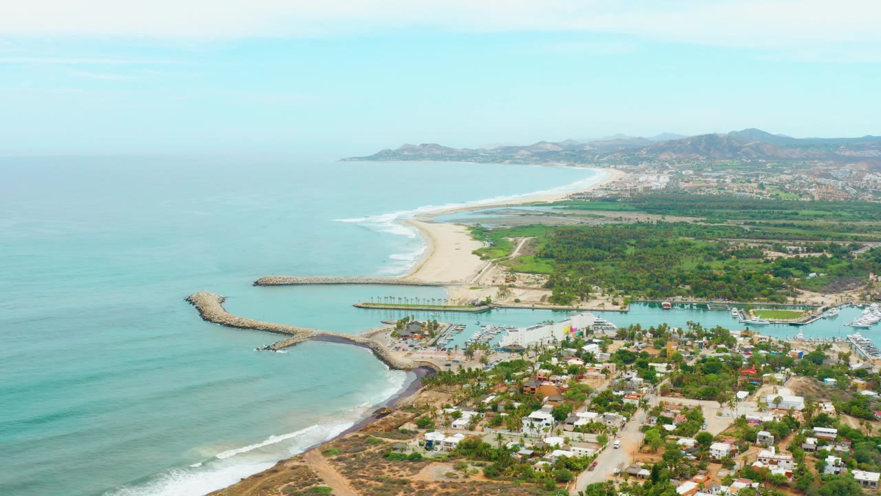 At 0:01, the La Playita Los Cabos aerial view keeps the harbor mouth and breakwater prominent while waves trace the beach and no visible people appear in the town or shoreline.