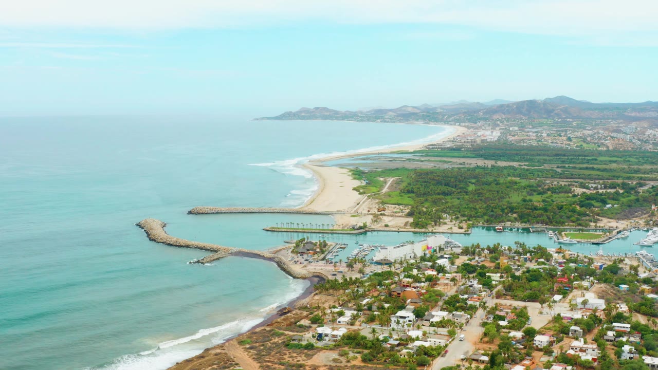At 0:02, the frame shows more open water on the left, the sandy beach curving into the distance, the harbor below, low buildings, green vegetation, hills, and no visible people.