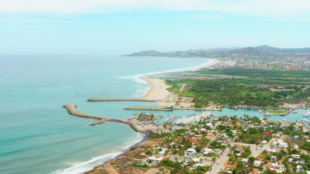 Coastal aerial with ocean foreground, harbor, town buildings, beach, and hills.