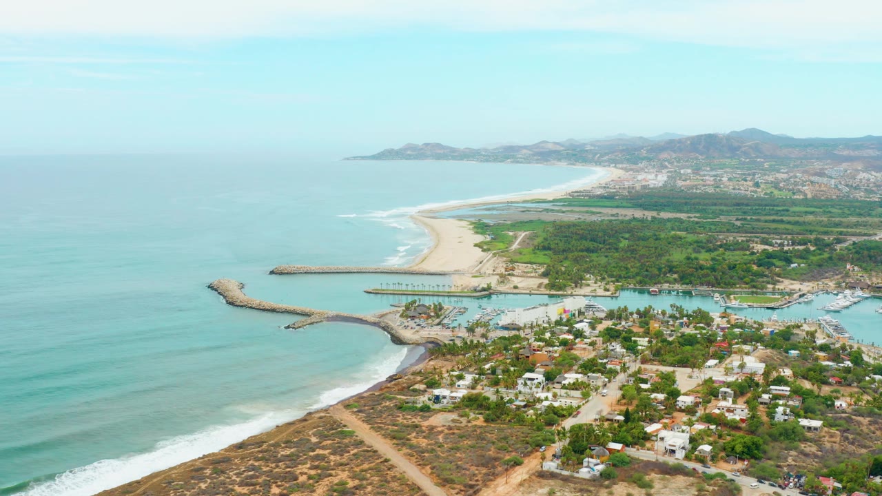 Drone view of blue ocean, curving beach, breakwater, harbor, and coastal homes.