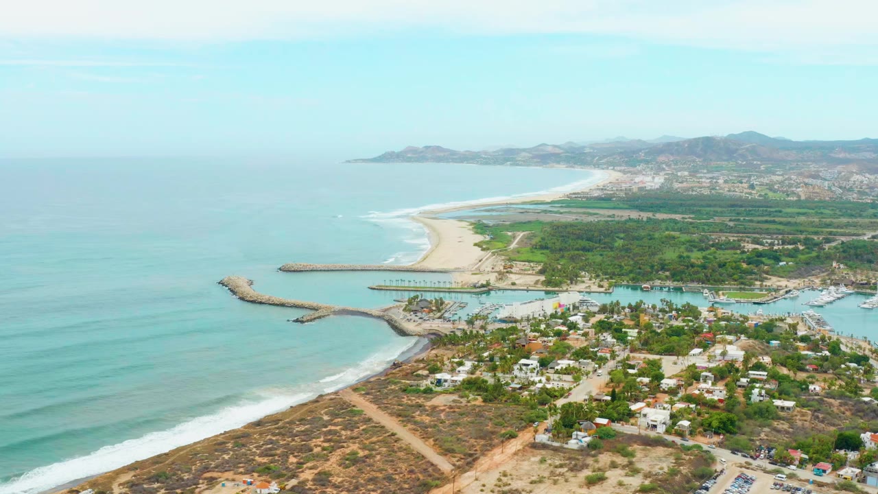Opening aerial frame over La Playita Los Cabos, Mexico, with turquoise ocean at left, a curving sandy beach, a small inlet, low buildings, green vegetation, distant hills, and no visible people.