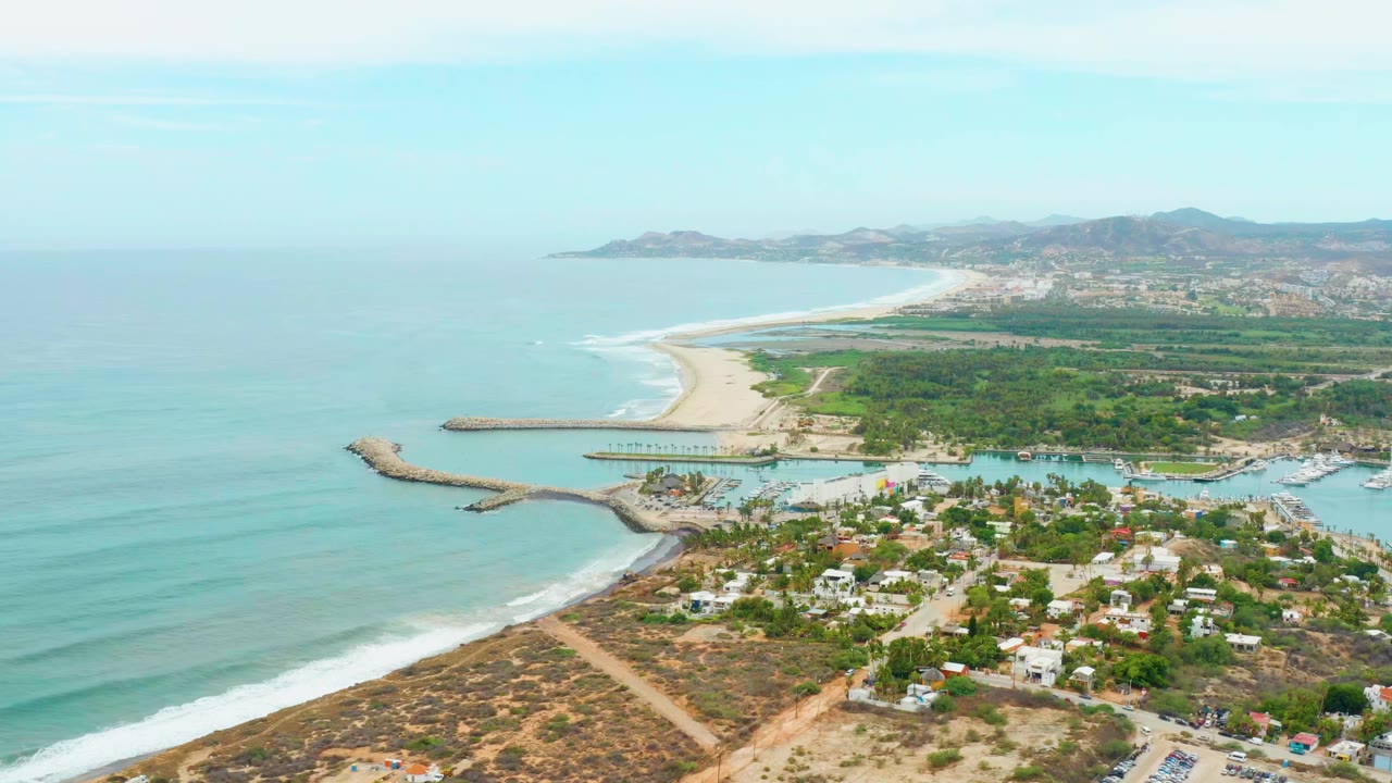 Aerial view at 0:01 over La Playita Los Cabos, Mexico, holding the ocean and surf on the left while the beach, inlet, clustered buildings, vegetation, and hazy hills remain visible with no visible people.