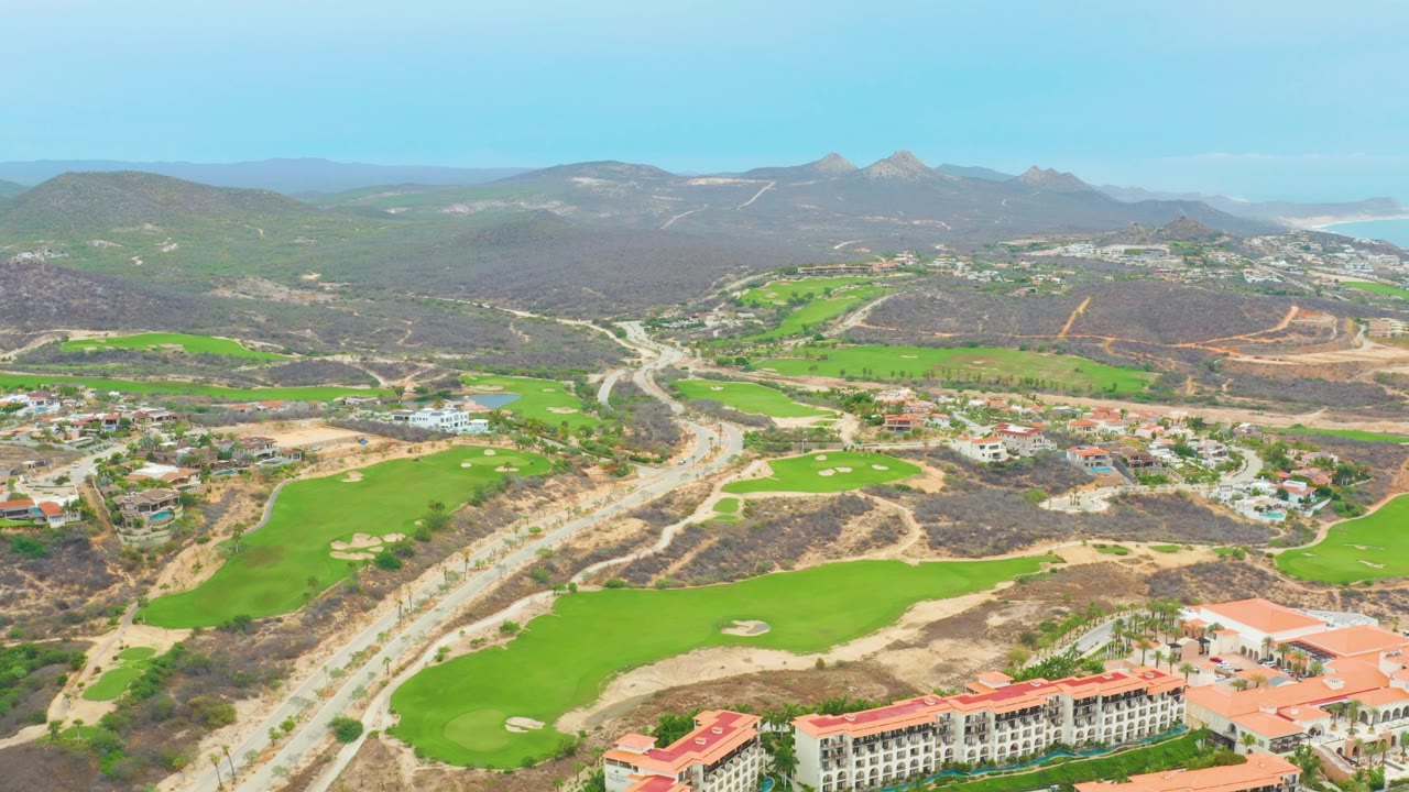 Wide drone view of green fairways, sandy terrain, residential buildings, roads, and mountains.