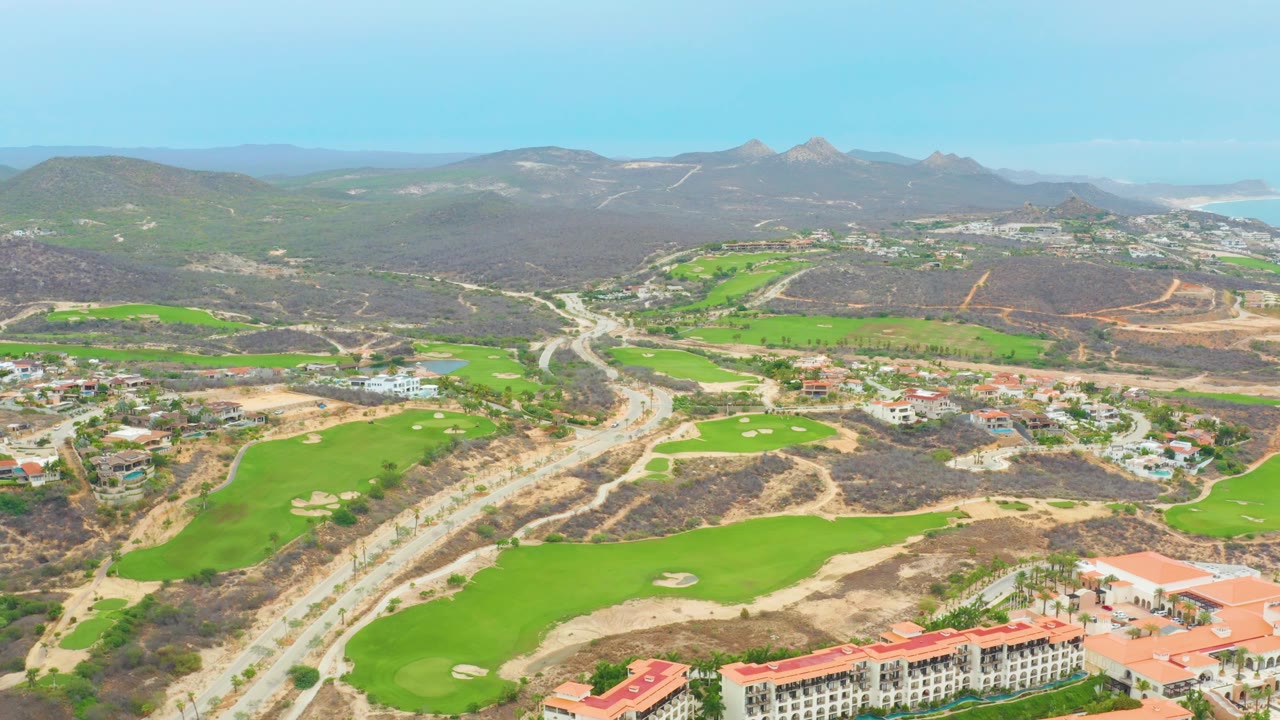 Drone aerial over La Playita Los Cabos showing bright green golf holes, pale sand traps, winding roads, scattered homes, arid hills, distant mountains, and no visible people.