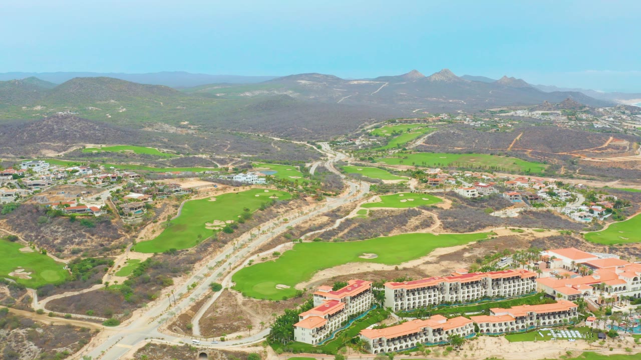 Wide aerial frame in La Playita Los Cabos showing green fairways beside graded dirt roads, resort buildings with red roofs along the lower edge, mountains on the horizon, and no visible people.
