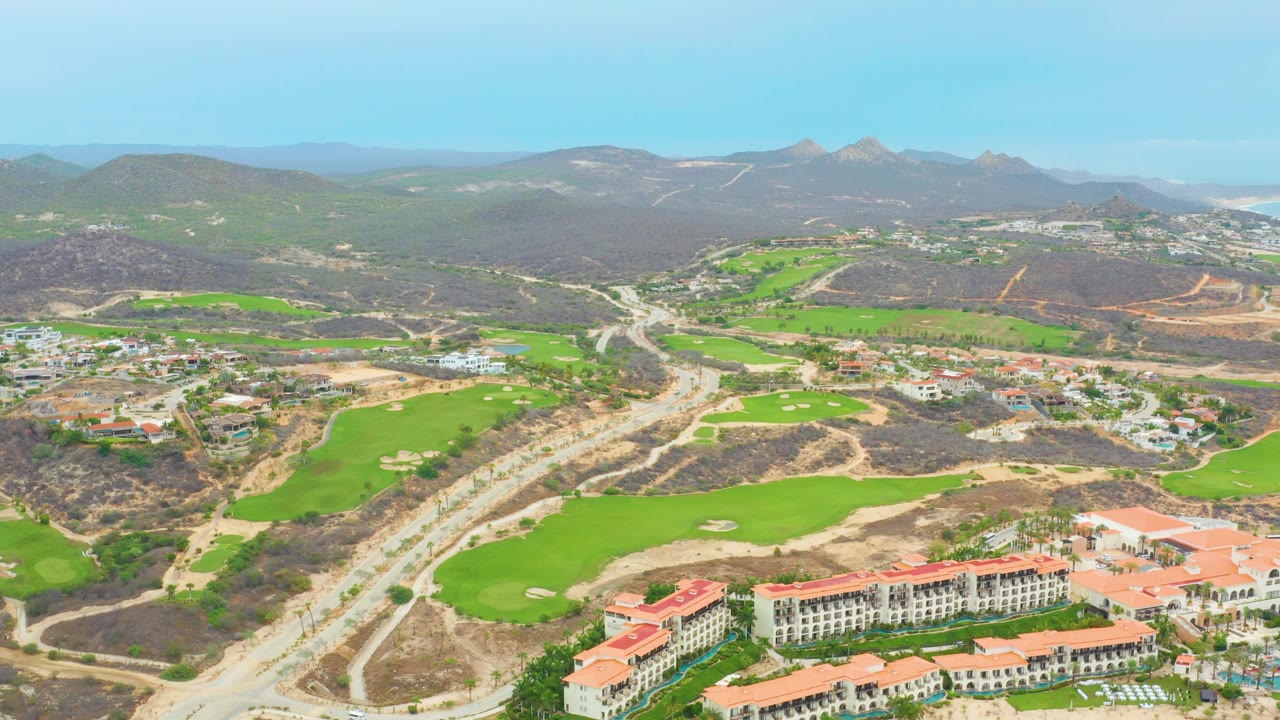 La Playita Los Cabos aerial with winding road construction through the center, golf fairways on both sides, resort buildings near the lower right, mountains in the distance, and no visible people.