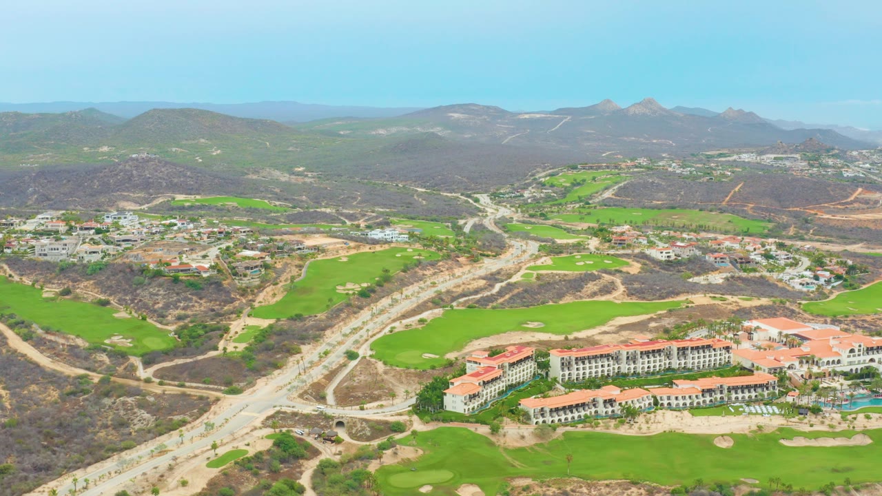 A wide aerial frame over La Playita Los Cabos shows a road cutting through dry hills beside bright golf greens, red-roofed resort buildings, distant mountains, and no visible people.