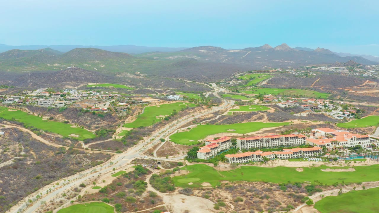 Aerial frame from La Playita Los Cabos showing a winding road, bright golf course greens, a red-roofed resort complex, dry hills, distant mountains, clear sky, and no visible people.