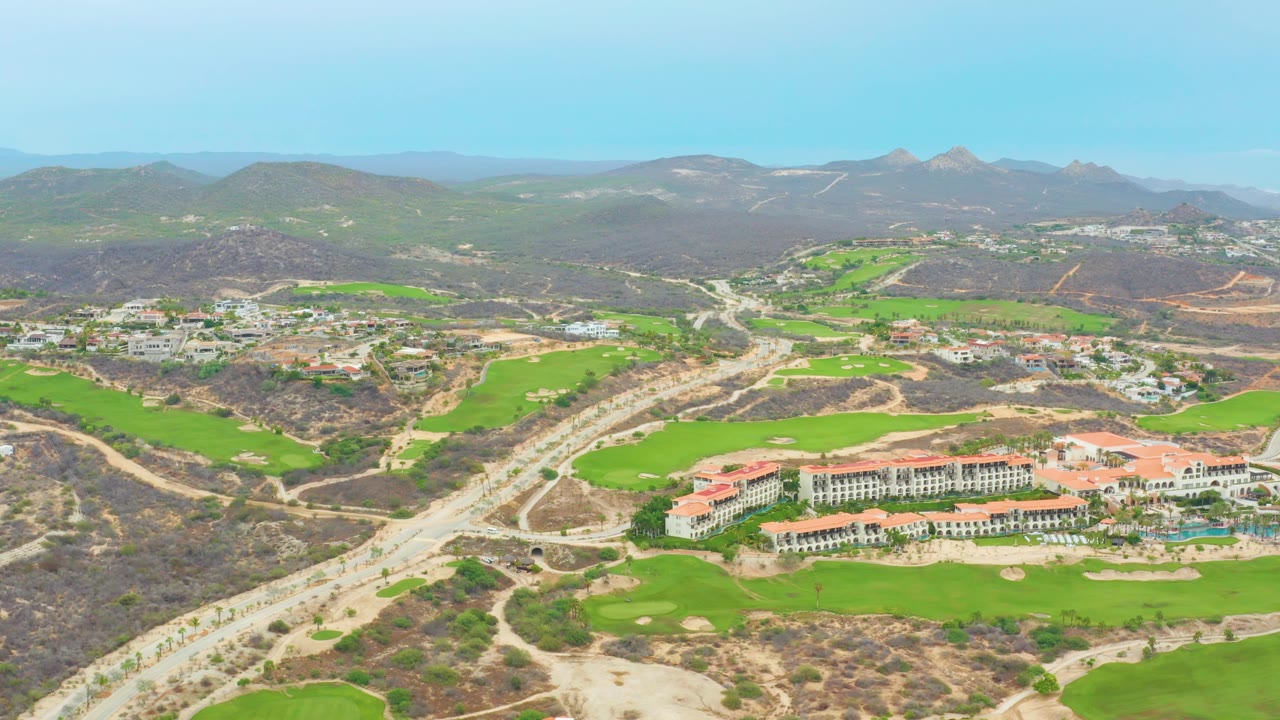 La Playita Los Cabos aerial view with the road crossing the left side of frame, green fairways and sand traps around red-roofed buildings, distant hills, and no visible people.