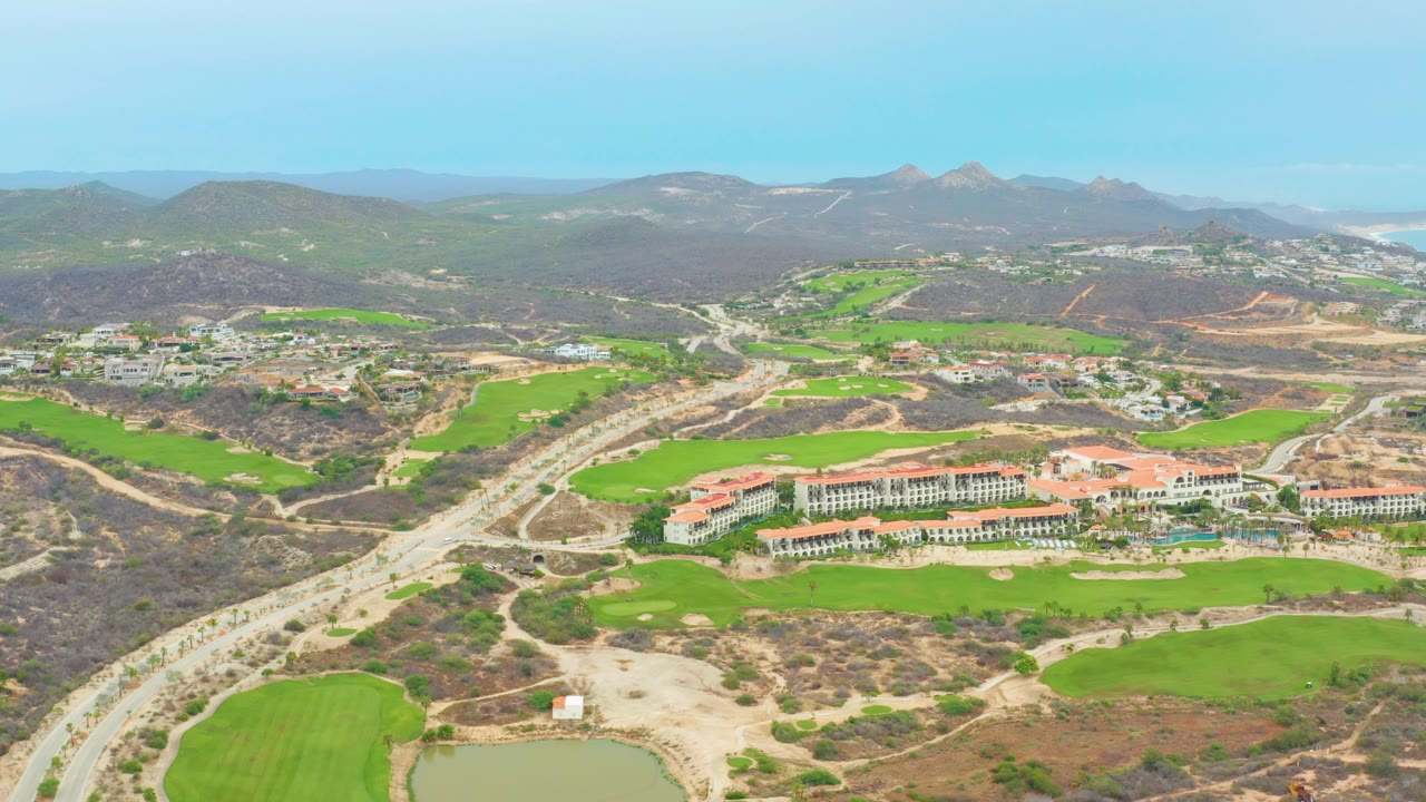 Opening aerial frame over La Playita Los Cabos, Mexico, showing green golf fairways beside a highway, low buildings, dry hills, distant mountains, clear sky, and no visible people.