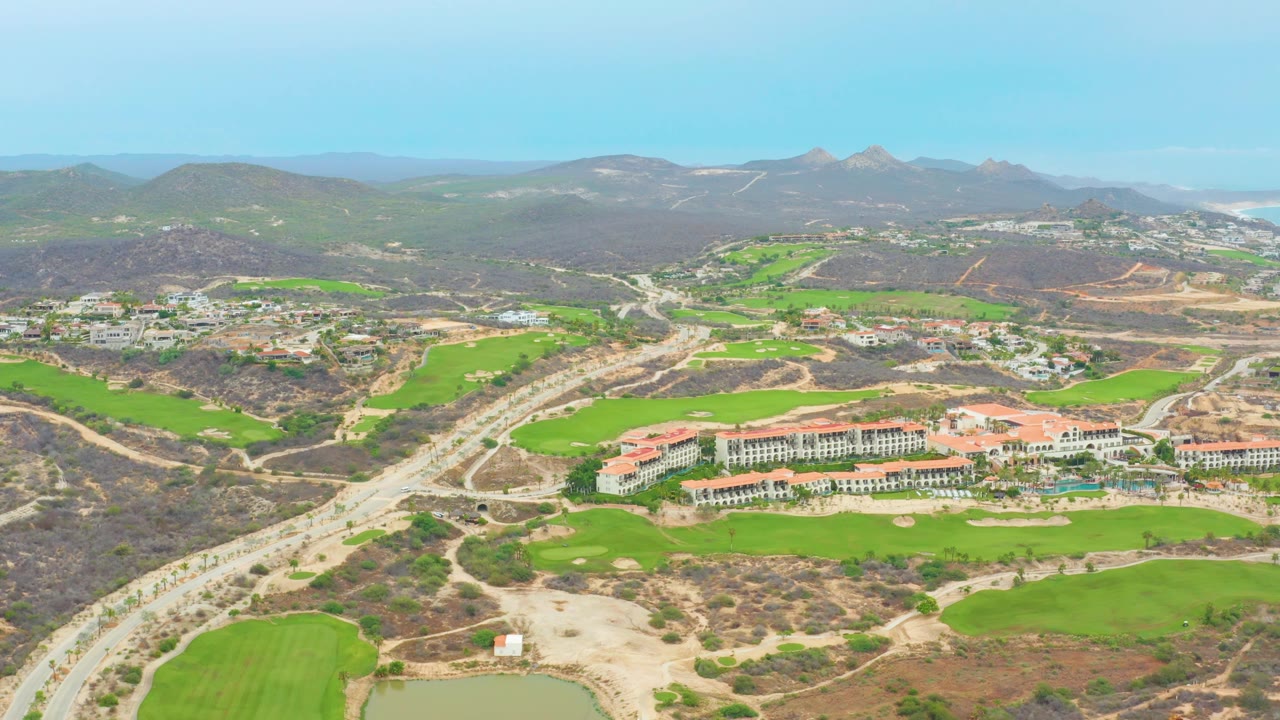 High aerial view over La Playita Los Cabos showing red-roofed buildings below the horizon, green golf holes, pale roads, dry open land, distant mountains, and no visible people.