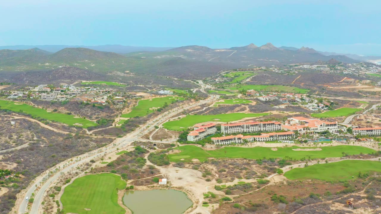 Opening aerial frame over La Playita Los Cabos with a winding road at left, green fields and a small pond below, red-roofed buildings in the midground, distant mountains, and no visible people.