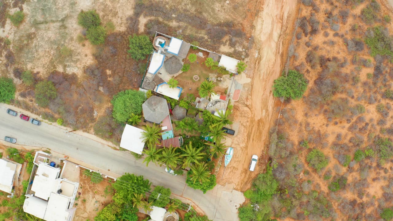 Aerial frame from La Playita Los Cabos with the road crossing the lower scene, a white vehicle on the dirt track, clustered buildings and palms, dry rocky ground, and no visible people.