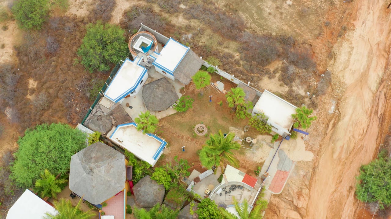 Aerial still of La Playita Los Cabos showing a compact group of roofs, palms in the courtyard, scrubby brown land around the property, and no visible people.