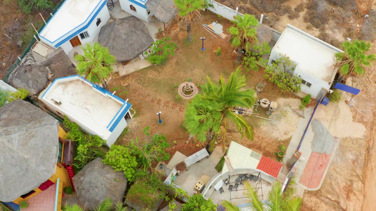 Overhead view of a La Playita Los Cabos courtyard with white buildings, blue trim, palm trees, thatched roofs, dirt ground, and no visible people.