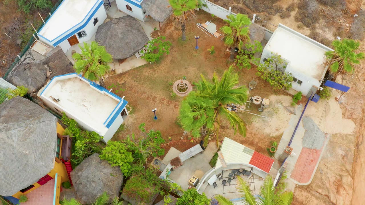 Aerial frame over La Playita Los Cabos showing white courtyard buildings, blue roof trim, a central dirt area, palms, thatched roofing, and no visible people.