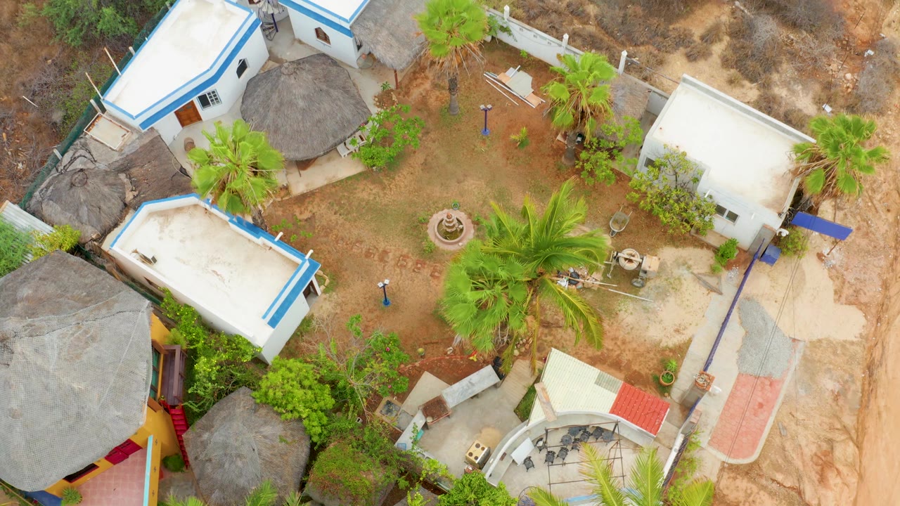 Drone-style overhead view in La Playita Los Cabos with thatched roof structures at left, white buildings around a dirt courtyard, dry land beyond, and no visible people.