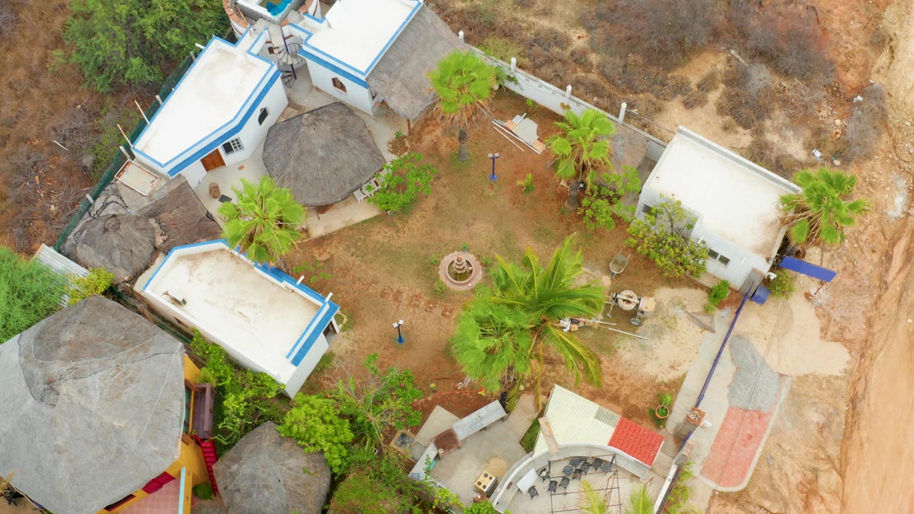 Top-down courtyard with white buildings, palms, and sandy road edge.
