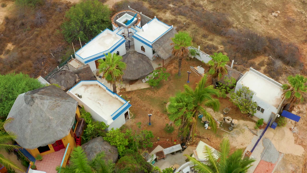 Aerial view in La Playita Los Cabos with a white flat-roof building near center, blue trim, thatched huts below it, palms, dry brushland, and no visible people.