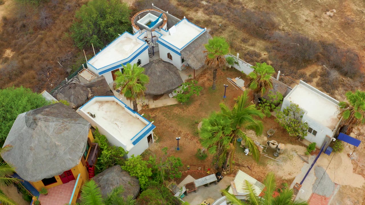 Drone-style overhead frame from La Playita Los Cabos showing the white buildings shifted slightly across the frame, with thatched roofs, palms, dry soil, brushland, and no visible people.