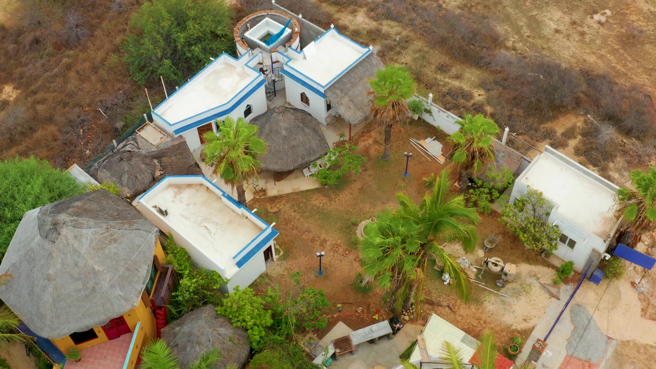 Closer overhead view of a large thatched roof beside white buildings and palms.