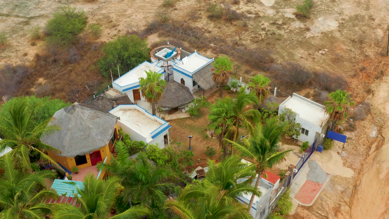 Opening overhead frame of a La Playita Los Cabos homestead with white buildings, blue trim, thatched roofs, palm trees, dry scrubland, and no visible people.
