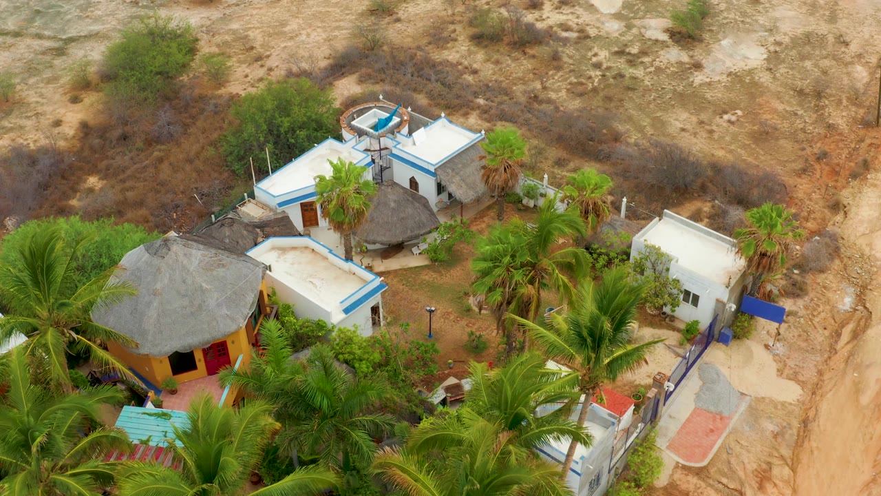 Second frame over La Playita Los Cabos shows the white-roofed compound and palapa roofs slightly farther right in frame, with palms below and no visible people.