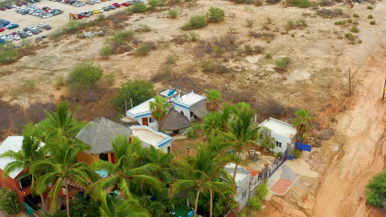 Aerial frame over La Playita Los Cabos showing palm trees around white and blue-accented buildings, a sandy dirt road, parked cars in the distant corner, and no visible people.