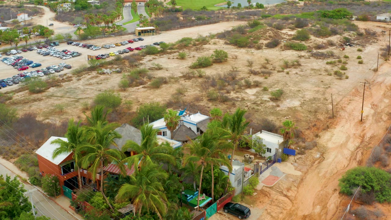 Opening aerial frame over La Playita Los Cabos showing palm-covered small buildings at the bottom, a sandy dirt road to the right, dry scrubland across the center, a parking lot at upper left, and no visible people.